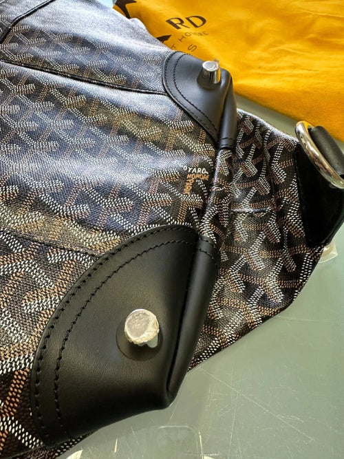 A close-up of a Goyard handbag featuring the brand's signature chevron monogram canvas in black, gray, and beige, with black leather trim and silver-tone hardware. A yellow Goyard dust bag is visible in the background.