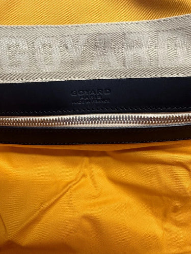 Close-up of a yellow garment with a wide beige woven band featuring embossed GOYARD lettering, a black leather trim, and a silver zipper. GOYARD PARIS branding is visible on the black section.