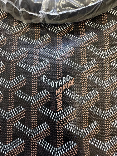Close-up of a Goyard monogram patterned material with the brand name 'Goyard Honore Se Goyard Paris' printed on it, with a crumpled clear plastic wrap visible at the top.