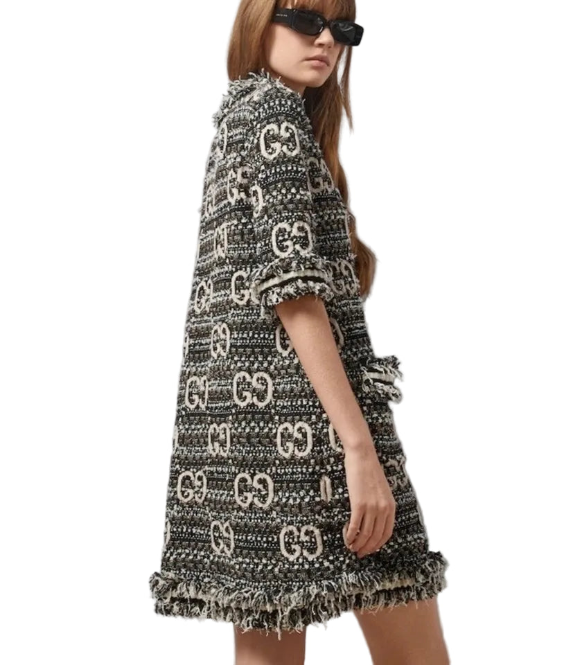 A model wearing a black and beige Gucci tweed mini dress with a fringed hem and an allover GG monogram pattern, styled with black sunglasses against a light gray background.