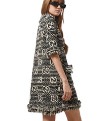 A model wearing a black and beige Gucci tweed mini dress with a fringed hem and an allover GG monogram pattern, styled with black sunglasses against a light gray background.
