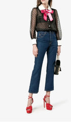 Woman wearing a black sheer top with a pink bow, blue jeans, and red high heels on a white background.