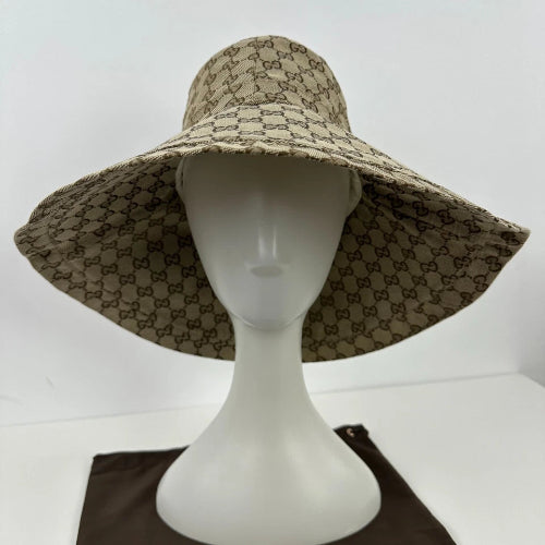 A wide-brimmed sun hat with a beige and brown repeating monogram pattern, displayed on a white mannequin head. The hat features a Gucci-style GG monogram pattern, and a dark brown fabric pouch is visible at the bottom.