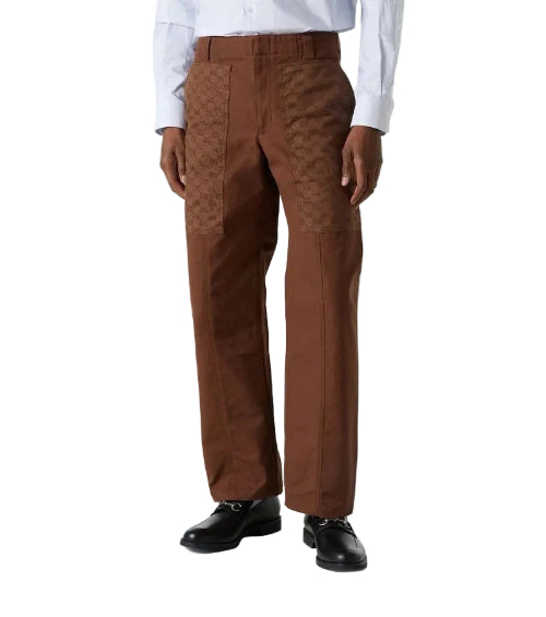 A person wearing brown tailored trousers with a subtle tonal patterned texture on the upper thighs, styled with a white dress shirt and black dress shoes against a light gray studio background.