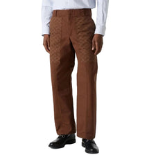A person wearing brown tailored trousers with a subtle tonal patterned texture on the upper thighs, styled with a white dress shirt and black dress shoes against a light gray studio background.