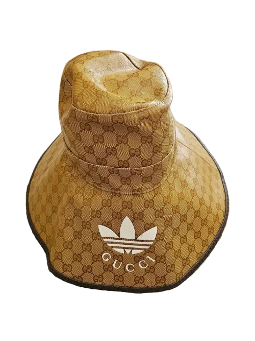 A Gucci x Adidas bucket hat in the signature brown GG monogram canvas, featuring a white Adidas trefoil logo and the word 'GUCCI' on the front, resting on a beige carpet.