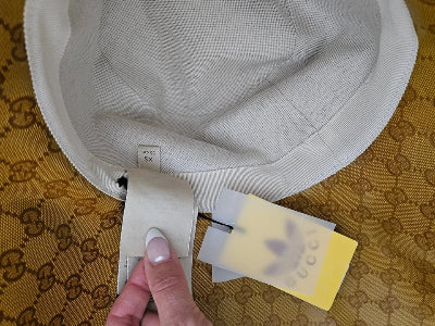 A close-up of a light beige fabric cap or bag interior with a hand holding a beige strap next to a yellow and white tag, placed on a gold and brown GG Supreme patterned surface.