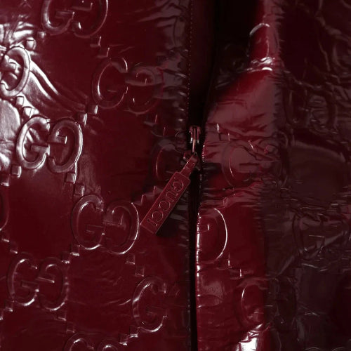 Close-up of a glossy burgundy quilted jacket featuring an embossed Gucci GG monogram pattern and a matching zipper pull with the word 'GUCCI'.