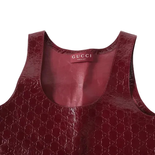 Close-up of a Gucci sleeveless top in glossy embossed GG Supreme leather in a deep burgundy red, showing the inner neck label with 'GUCCI MILAN'.