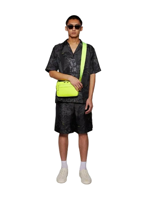 Person wearing a black patterned shirt and shorts with a neon green bag on a white background