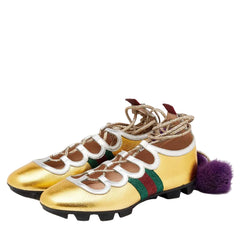 A pair of metallic gold high-top sneakers with brown suede overlays, white trim, a green and red web stripe, beige laces, and a purple pom-pom on a light gray background.