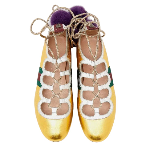 A pair of metallic gold Gucci ballet flats with white cutout straps, green and red web stripes, gold lace-up detailing, and purple faux-fur pom-poms on a light gray background.