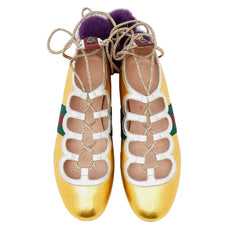 A pair of metallic gold Gucci ballet flats with white cutout straps, green and red web stripes, gold lace-up detailing, and purple faux-fur pom-poms on a light gray background.