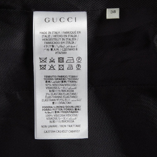 A close-up of a white Gucci garment care label sewn onto black textured fabric, with the Gucci brand name clearly printed at the top and a small size marker reading 38 nearby.