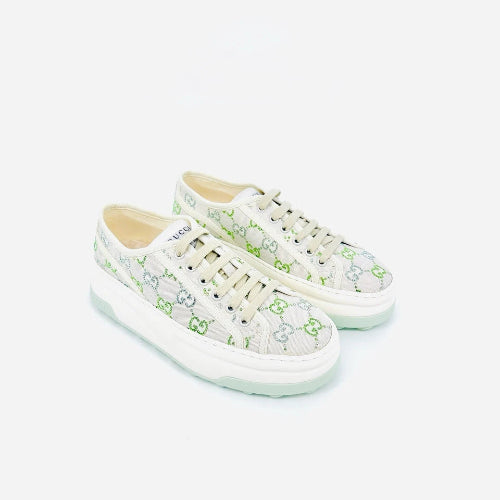 A pair of Gucci low-top sneakers featuring a light beige canvas upper with a green GG monogram pattern, cream laces, and a thick white platform sole with a pale mint green edge, set against a plain light gray background.