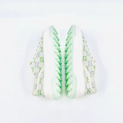 A pair of low-top sneakers shown from the side, featuring a white canvas upper with a green and gray monogram pattern, white laces, and a chunky white sole with a mint green textured zigzag design on the outsole.