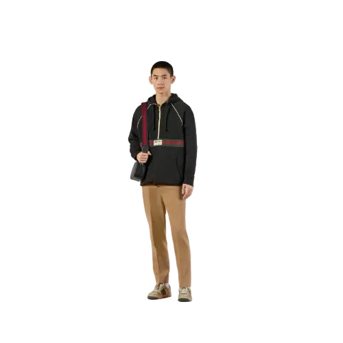 A male model stands in a studio wearing a black Gucci hoodie with the brand's signature red and green web stripe across the chest, tan trousers, and carrying a black shoulder bag.
