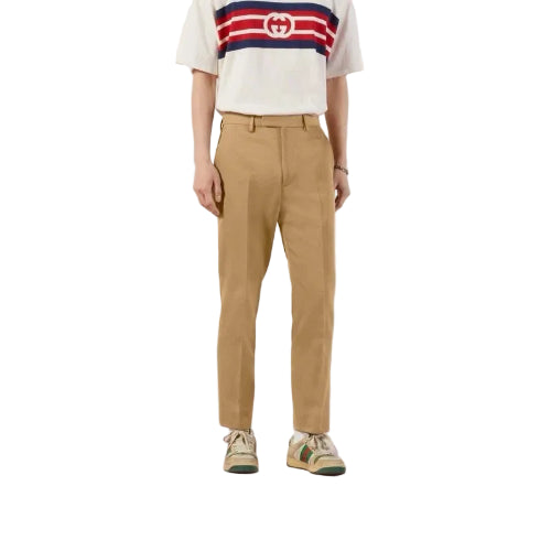 A model wearing tan chino pants, a white Gucci t-shirt with red and navy stripes, and green and beige sneakers, standing against a light pink and white studio background.