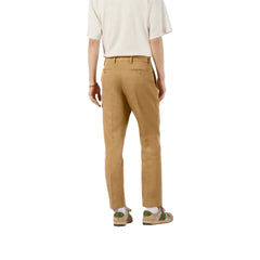 A person shown from the back wearing tan slim-fit trousers, a cream short-sleeve top, and green-and-beige loafers on a pale pink floor against a white wall.