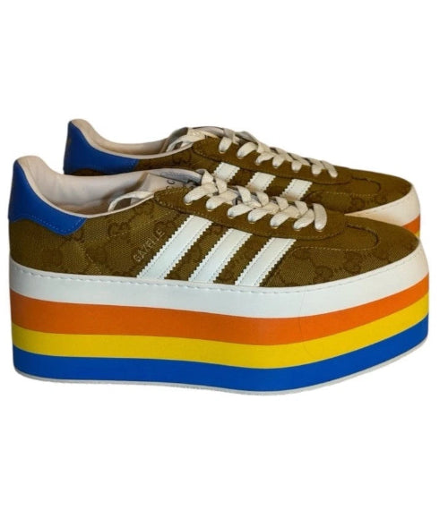 A pair of platform sneakers with a brown leather upper, white three-stripes branding, and a thick platform sole featuring a yellow, orange, and blue gradient.