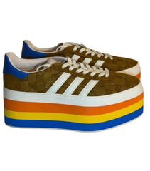 A pair of platform sneakers with a brown leather upper, white three-stripes branding, and a thick platform sole featuring a yellow, orange, and blue gradient.