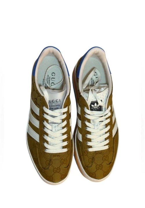 A pair of Gucci GG Marmont low-top sneakers in olive brown suede with white laces, white side stripes, and blue accents on the collar and heel, shown from a top-down view on a white background.