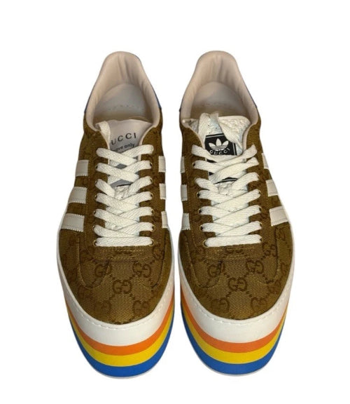 A pair of Gucci x Adidas low-top sneakers featuring brown GG Supreme canvas, white laces, white side stripes, and a distinctive rainbow-striped platform sole.