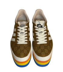 A pair of Gucci x Adidas low-top sneakers featuring brown GG Supreme canvas, white laces, white side stripes, and a distinctive rainbow-striped platform sole.