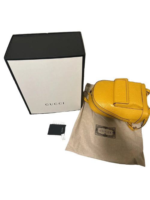 Yellow Gucci handbag on a beige cloth with a black and white Gucci box and small black item on a gray carpeted floor.