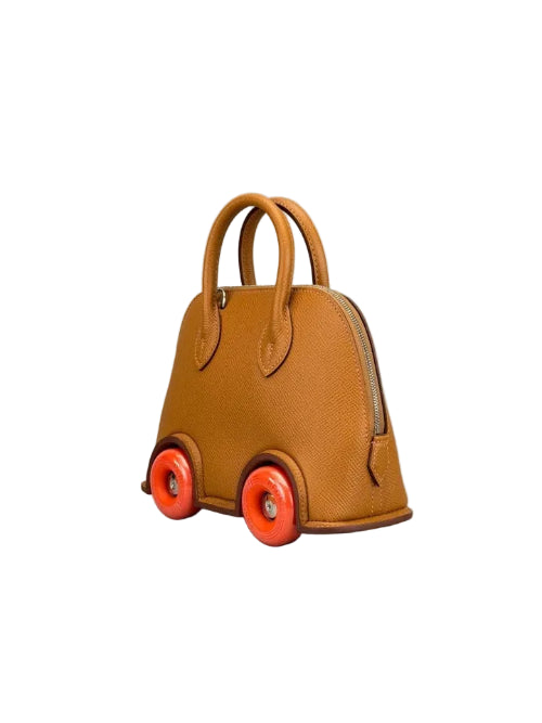 A small structured handbag designed like a toy car, made of tan textured leather with bright orange wheels and gold-tone hardware, photographed on a neutral gray studio background.