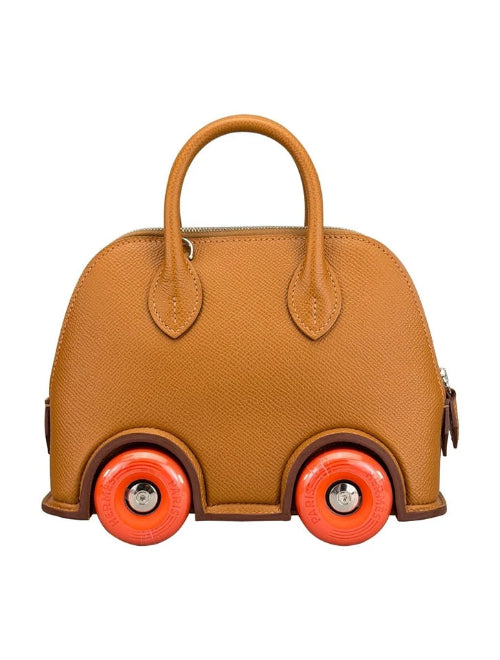 A structured handbag designed to look like a toy car, made of textured tan leather with bright orange wheels and silver metal wheel hubs, shown against a plain white background.
