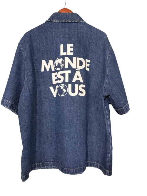 Blue denim shirt with 'Le Monde Est A Vous' text on a textured wall background
