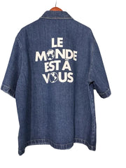 Blue denim shirt with 'Le Monde Est A Vous' text on a textured wall background
