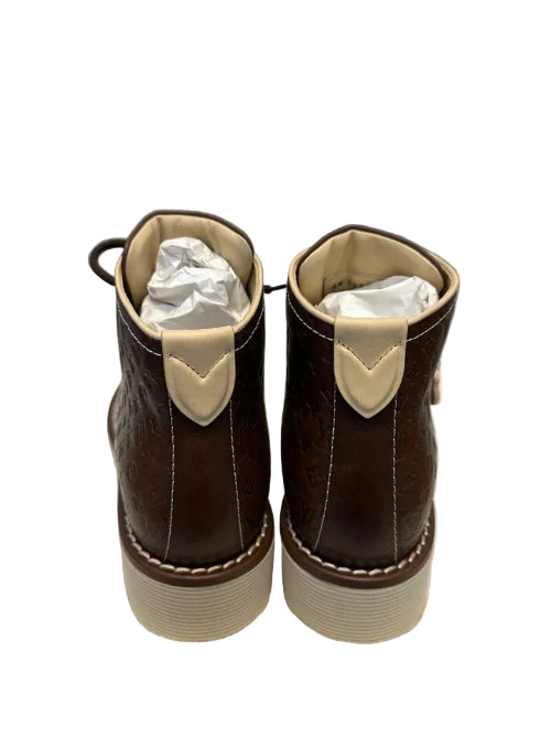 A pair of dark brown leather boots with white trim and white plastic inserts, resting on a white textured surface against a light gray wall.