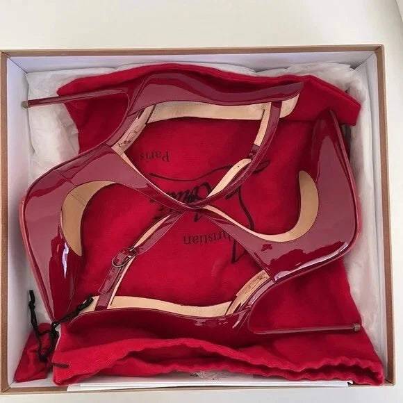 A pair of glossy red patent leather high-heeled shoes with beige straps, presented inside a white box with a red dust bag featuring Christian Dior branding.