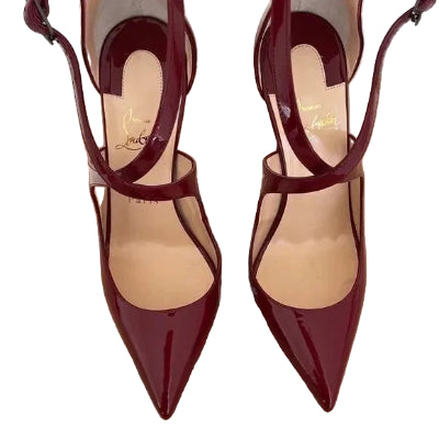 A pair of pointed-toe high heels in glossy deep burgundy patent leather with ankle straps, displayed on a brown shoebox. The brand name 'Christian Louboutin' is visible in gold script on the insoles.