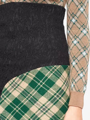 Close-up of a long-sleeve knit top with a color-blocked design: black textured lace on the upper section, green and beige tartan plaid on the lower section and left sleeve, and a beige plaid pattern on the right sleeve.