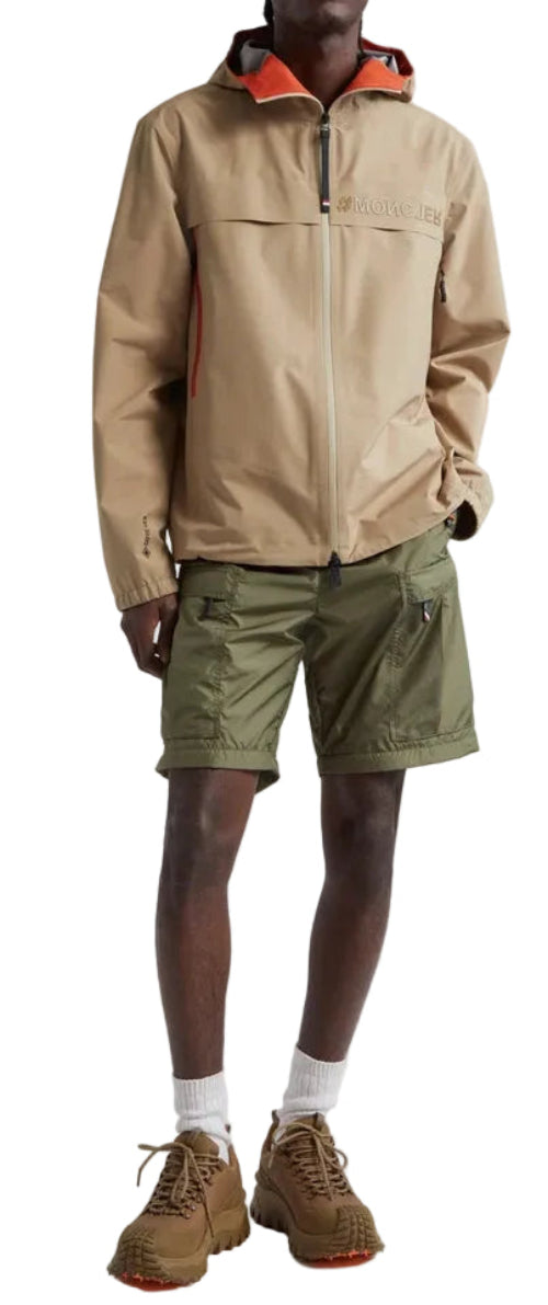 A man wearing a beige hooded jacket, olive green shorts, and brown boots, shown from the waist up against a plain light background.