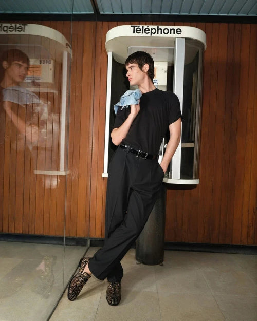 A model wearing a black t-shirt, black wide-leg trousers, and leopard-print loafers poses in front of a retro glass telephone booth with a wood-paneled wall.