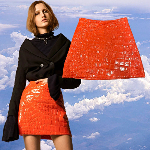 A model wears a bright orange crocodile-embossed mini skirt with a black double-breasted blazer, set against a sky and clouds background, with a larger flat view of the orange skirt shown to the right.