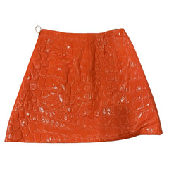 An orange crocodile-embossed faux-leather mini skirt with a glossy finish, shown against a sky and clouds background.