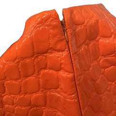 Close-up of a bright orange crocodile-embossed leather item being held by a hand, showing the glossy textured surface and a gold-toned zipper.