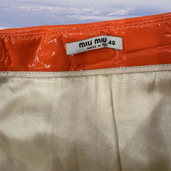 Close-up of a Miu Miu garment waistband showing a glossy orange patent leather trim and a cream-colored satin inner lining, with the brand label reading 'miu miu MADE IN MIU 40'.