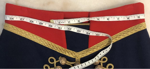 A close-up of a navy blue garment with a red waistband, gold braided trim, and a white measuring tape laid across the front to show dimensions.