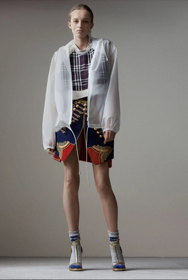 A model stands against a plain light gray studio background wearing a white long-sleeve shirt, a black-and-red patterned vest, black shorts, white socks, and black shoes.