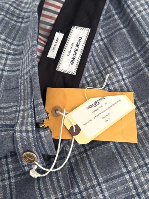 Close-up of a plaid blazer fabric in blue and white, showing a black interior lining with a white sewn-in label reading THOM BROWNE, plus an orange paper tag attached with white string.