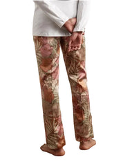 A person shown from the waist down wearing floral print trousers with a white long-sleeve top, standing barefoot on a wooden parquet floor against a plain wall.