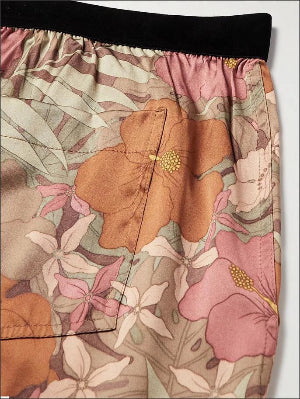 Close-up of a floral-patterned garment, likely shorts or a skirt, featuring large orange, pink, and white flowers with green leaves on a beige background, finished with a black elastic waistband.