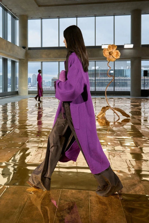 Woman in a purple coat walking in a modern building with large windows and a sculpture.