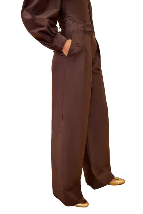 A model wearing dark brown wide-leg trousers with a matching top, standing on a patterned beige carpet against a textured cream wall.
